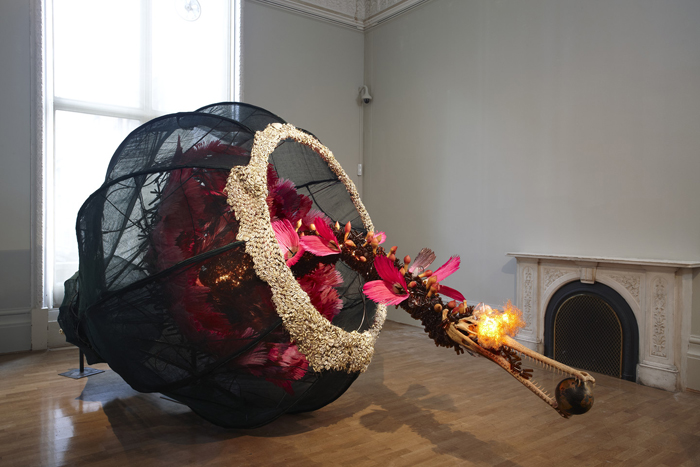 Rina Banerjee, The world as burnt fruit—When empires feuded for populations and plantations, buried in colonial and ancient currency a Gharial appeared from an inky melon—hot with blossom sprang forth to swallow the world not yet whole as burnt fruit, 2009. Fans, feather, cowrie shells, resin alligator, skull, globe, glass vials, light bulbs, gourds, steel wire and Japanese mosquito nets, 90 x 253 x 90 inches. Kiran Nadar Museum of Art, New Delhi, India.